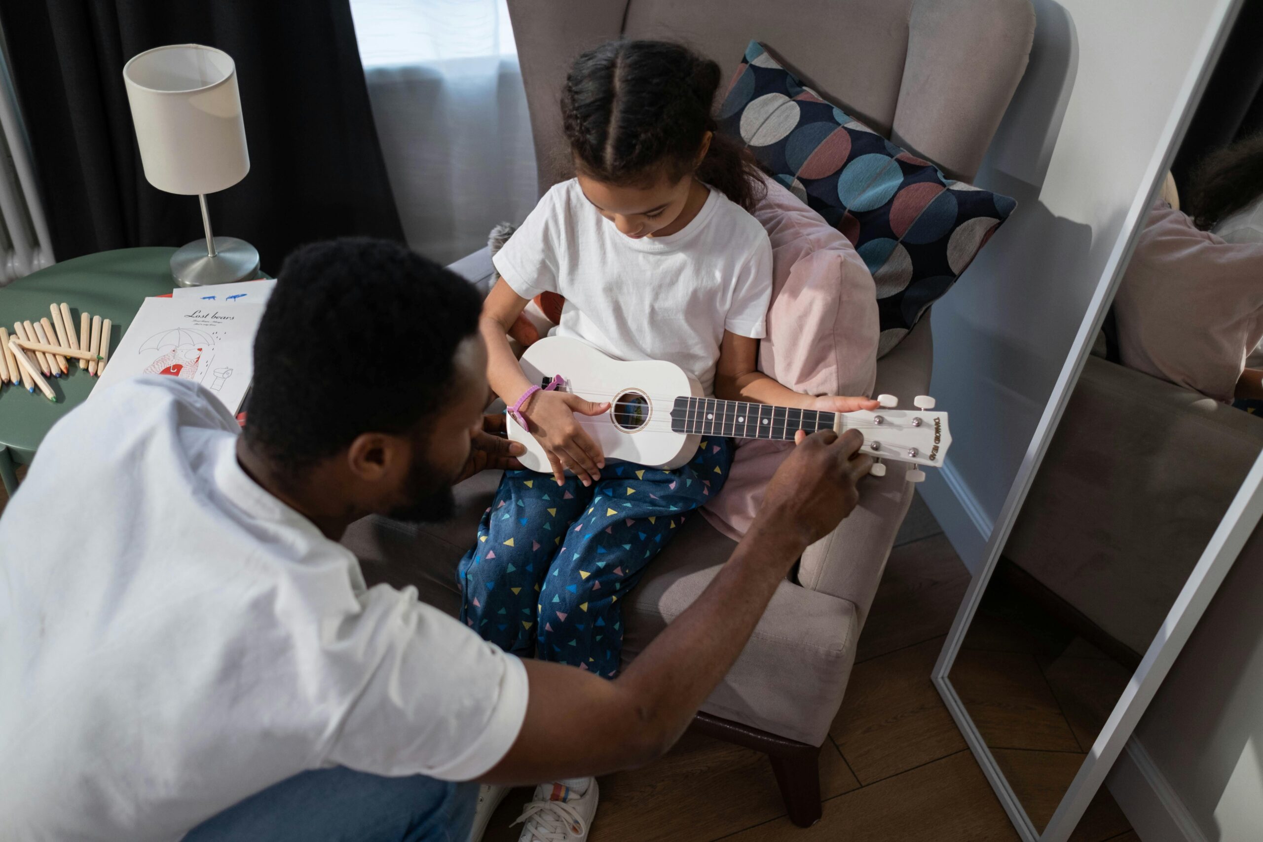 A man helps a young girl tune a white ukulele as she sits attentively on a chair.