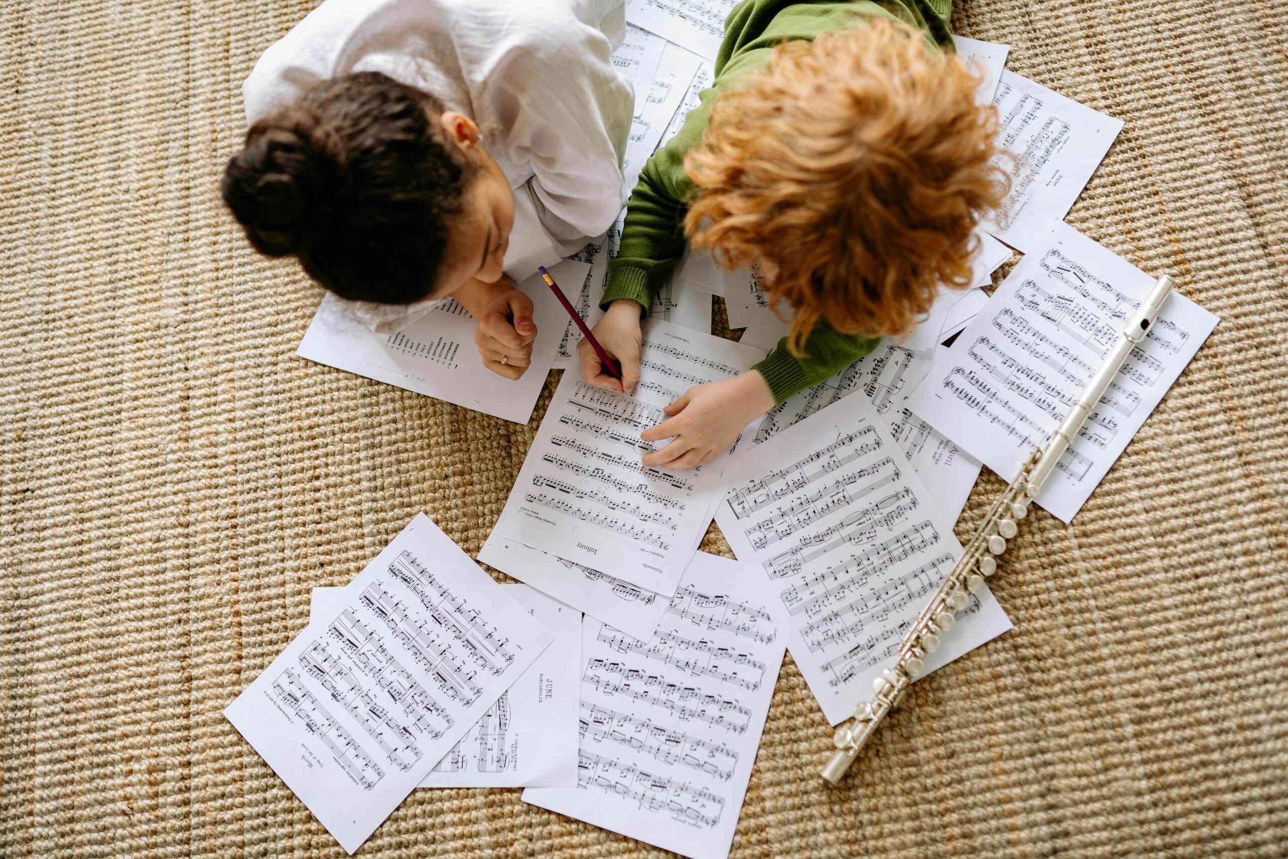 Two young students lie on the floor reviewing and annotating sheet music, surrounded by musical scores and a flute.