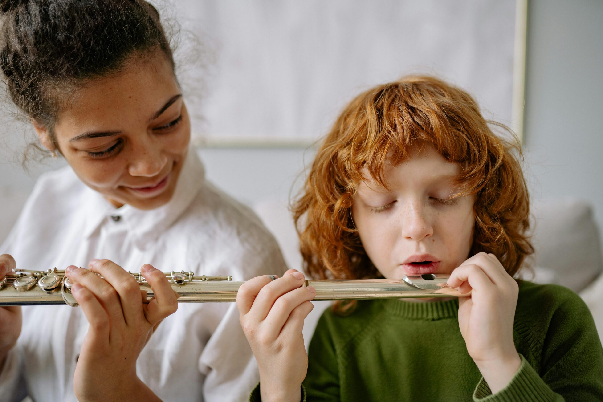 A young student practices embouchure technique on the flute with guidance from a smiling peer or instructor.