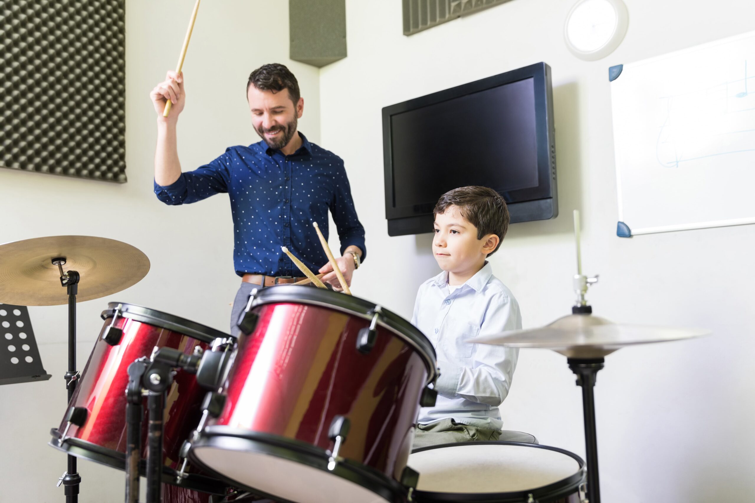 A drum teacher enthusiastically guides a young student through a lesson in a soundproofed music classroom.