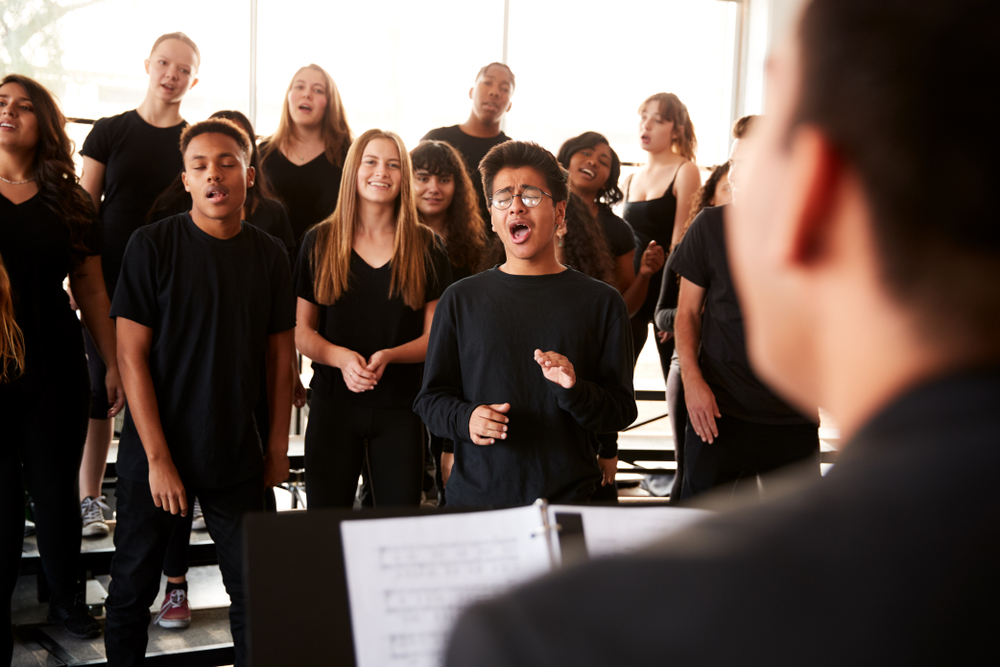 Male,And,Female,Students,Singing,In,Choir,With,Teacher,At A diverse group of students performs in a choral rehearsal, guided by a conductor in a well-lit music classroom / performing arts school.