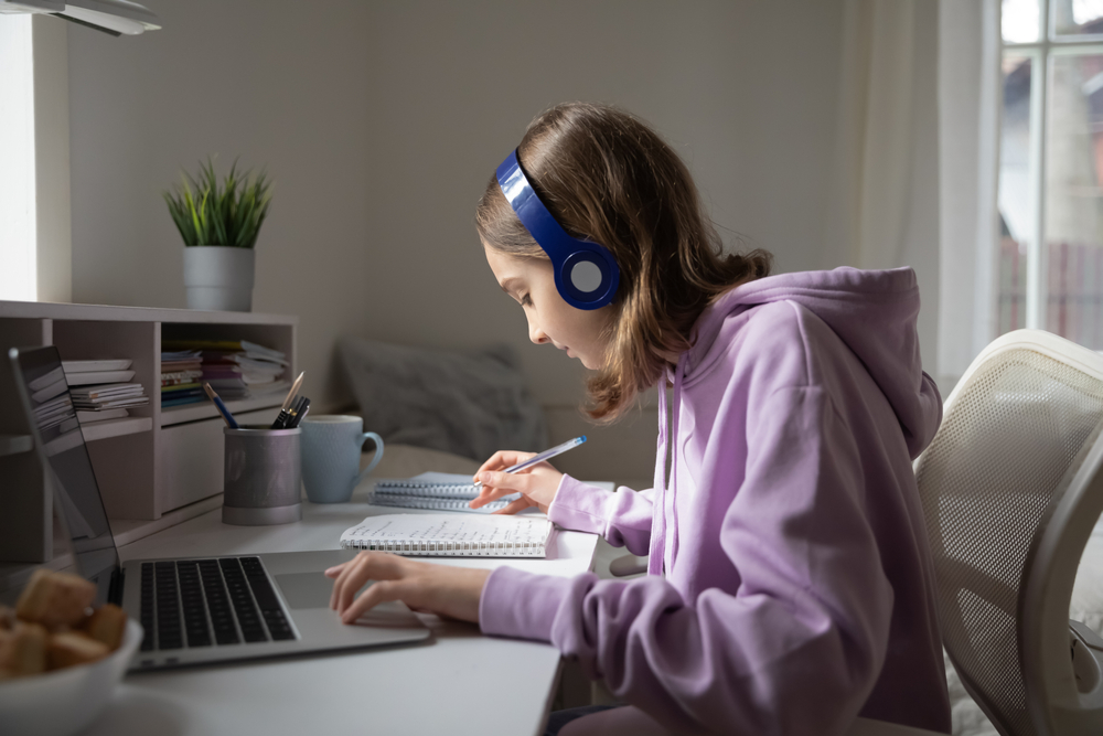 Teen,Girl,School,Pupil,Wearing,Headphones,Studying,Online,From,Home A focused student wears headphones while studying at a laptop, taking notes during an online lesson from home.