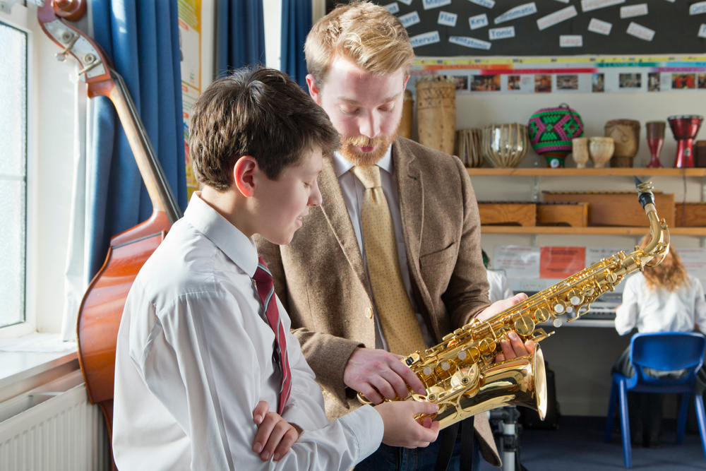 A music teacher provides one-on-one saxophone instruction to a student in a classroom filled with instruments.