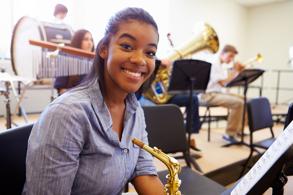 Female,Pupil,Playing,Saxophone,In,High,School,Orchestra Highschool girl smiling at the cameron while sitting in band class with her saxophone