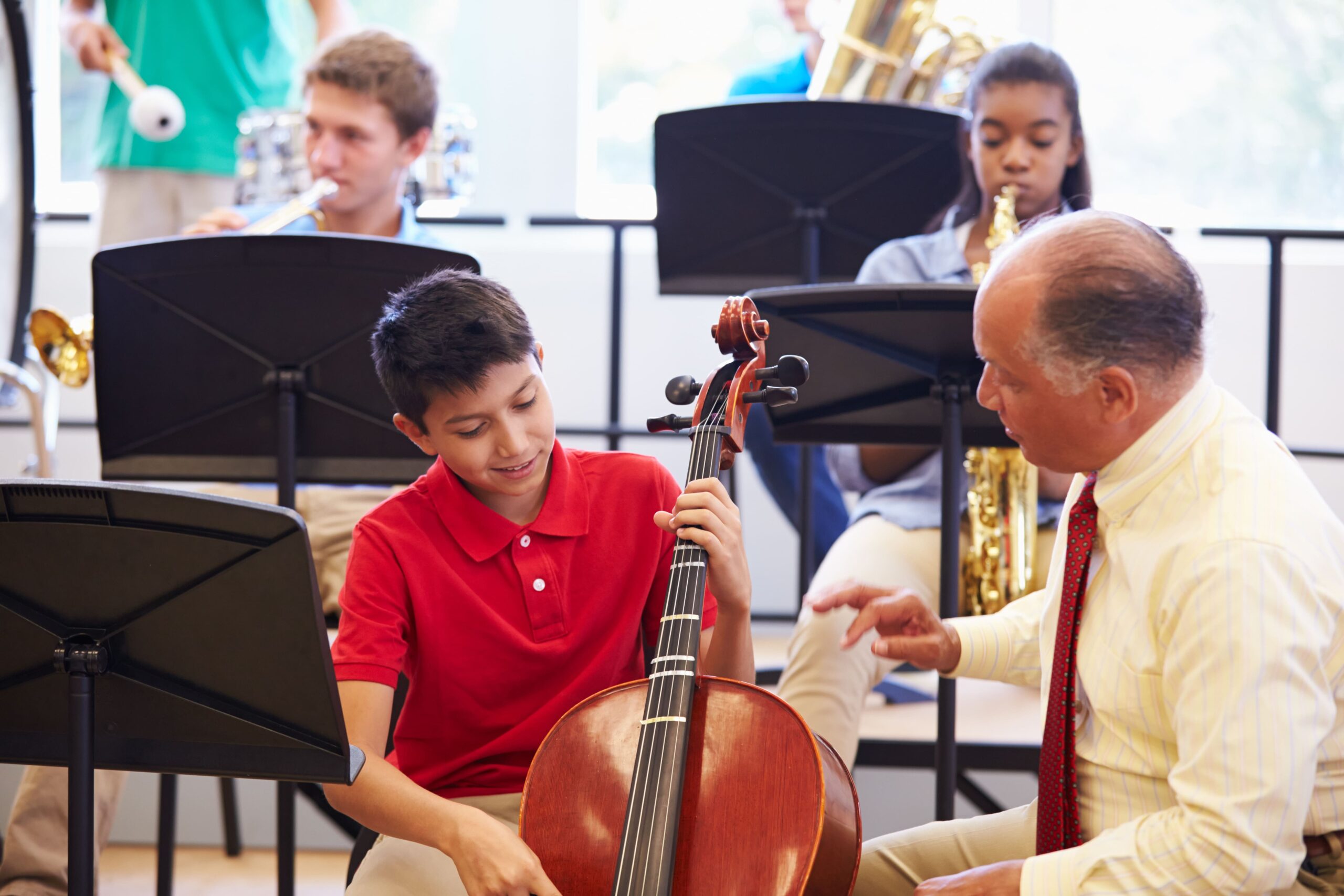 A music teacher provides instruction to a student holding a cello during a band class rehearsal.