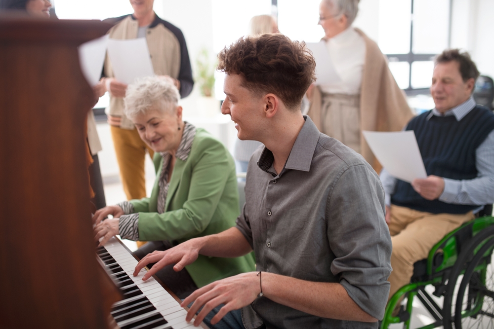 Senior,Woman,With,Young,Teacher,Playing,At,Piano,In,Choir Young man playing piano with elderly woman, smiling