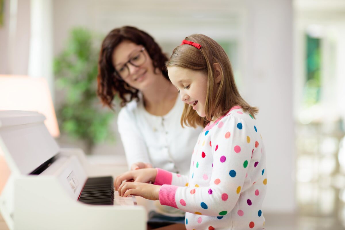 A young girl smiles while playing the piano as her teacher guides her hands on the keys.