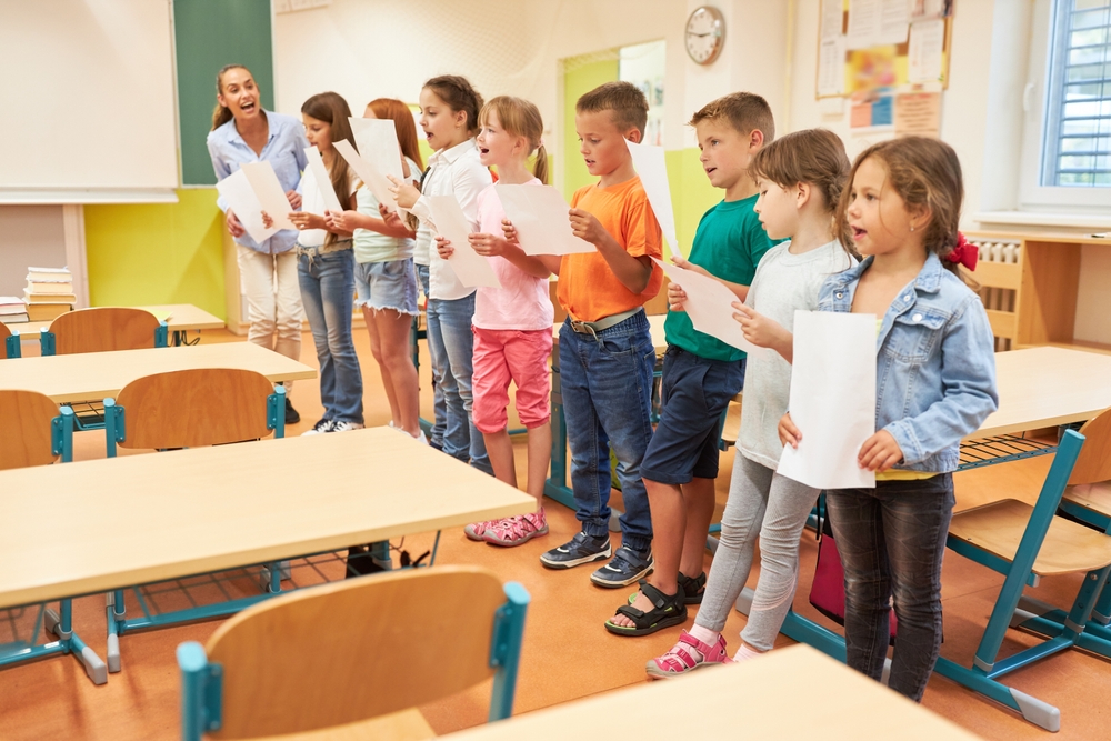 Group,Of,Elementary,School,Children,Singing,Song,During,Choir,Practice A group of elementary students practices singing in a classroom, holding lyric sheets and following their enthusiastic teacher.