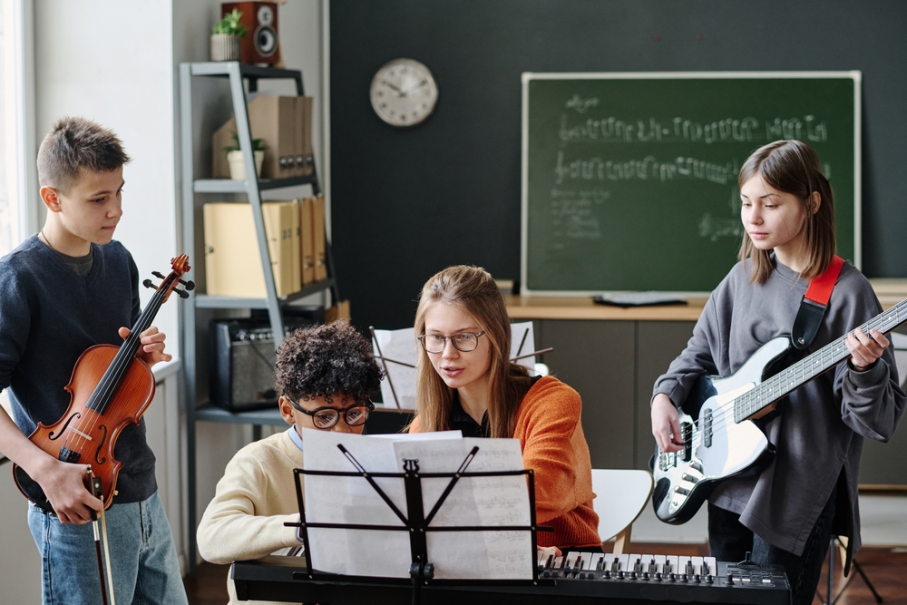 Medium,Long,Shot,Of,Young,Music,Teacher,Explaining,Her,Students Woman sitting at piano teaching three kids, two of them standing next to her and the other sitting on the bench