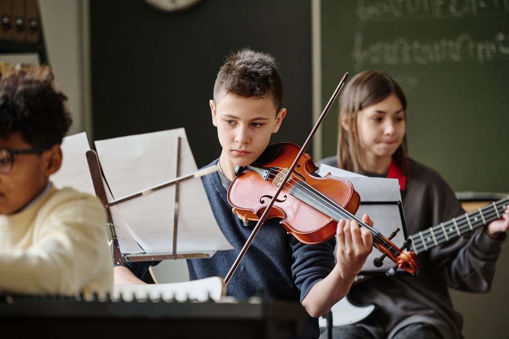 Selective,Focus,Of,Boy,Reading,Sheet,Music,And,Playing,Violin Middle school aged boy focusing on sheet music while playing violin in music class