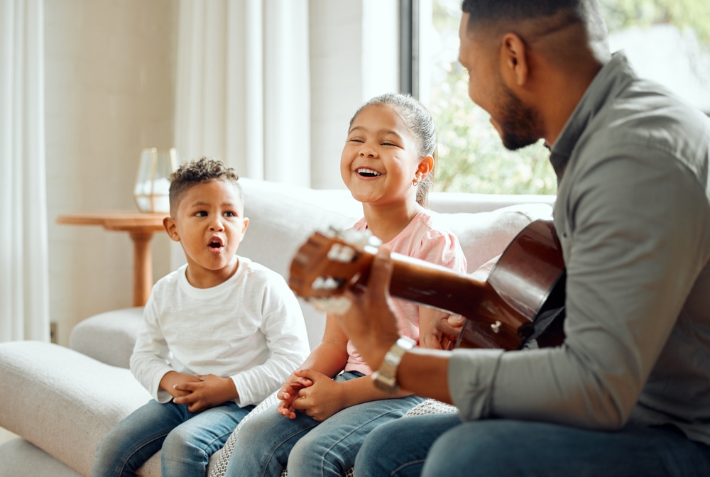 Singing,,Children,And,Dad,With,Guitar,,Living,Room,And,Teaching Two young children sing and laugh while their teacher plays guitar during a joyful music lesson at home.
