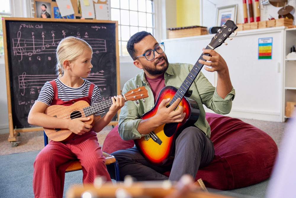 Teacher,Assisting,Kid,To,Play,Musical,Instrument,In,Classroom,At Man in glasses sitting on beanbag chair giving guitar lesson to young girl in a classroom