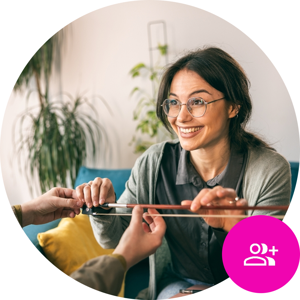 CRM Featured Image – Detail Female music teacher holding violin bow and looking at student across the table - CRM Icon in the bottom right of the screen on brand pink background